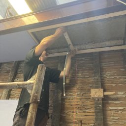Deen-Voic - Pracownik na rusztowaniu montuje drewniane listwy do sufitu z widoczną instalacją elektryczną, ściany w stanie surowym z widocznymi warstwami starej farby i tynku.