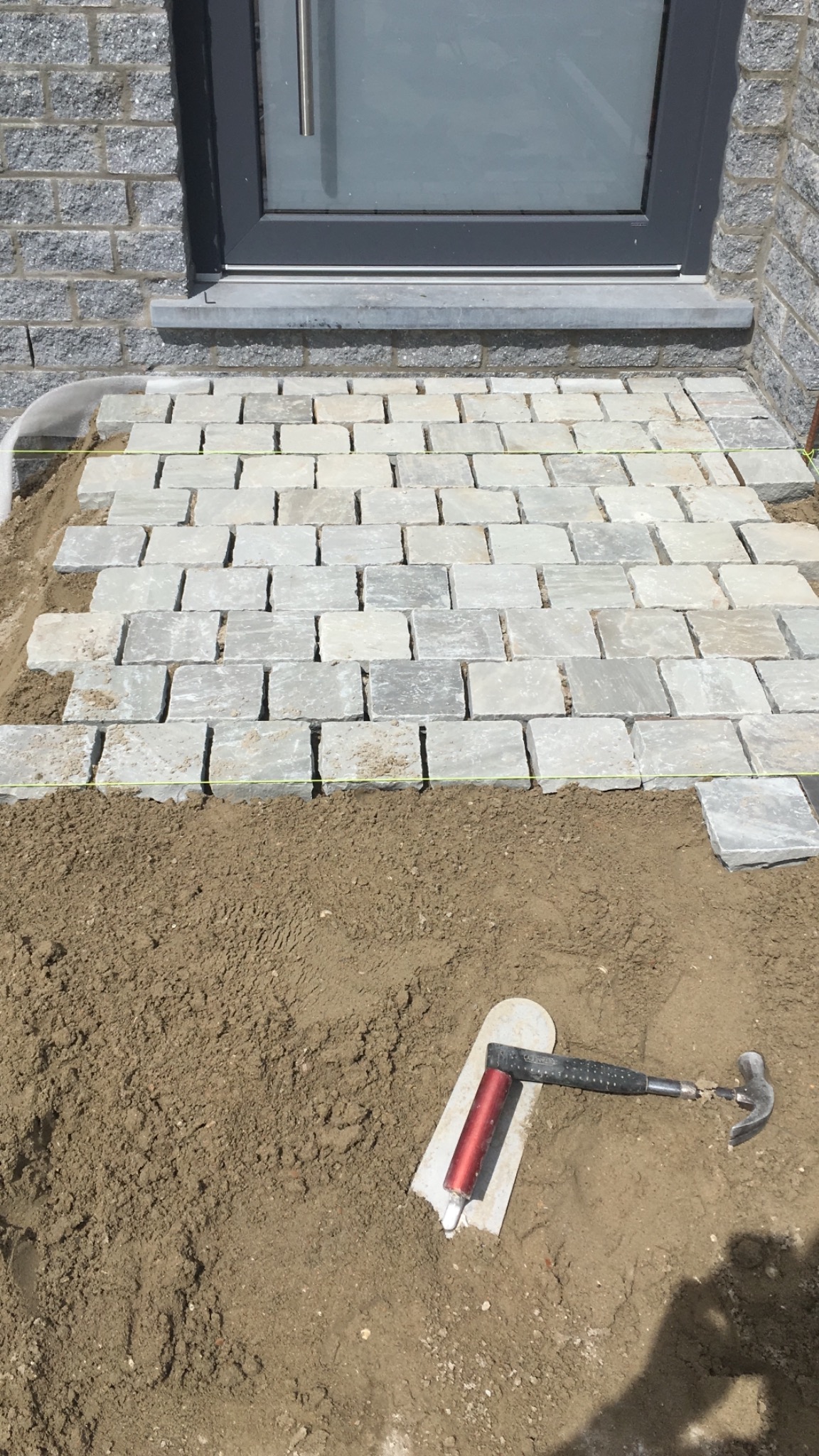 Układanie szarej kostki brukowej przed wejściem do budynku, widoczna poziomica i młotek na piasku.