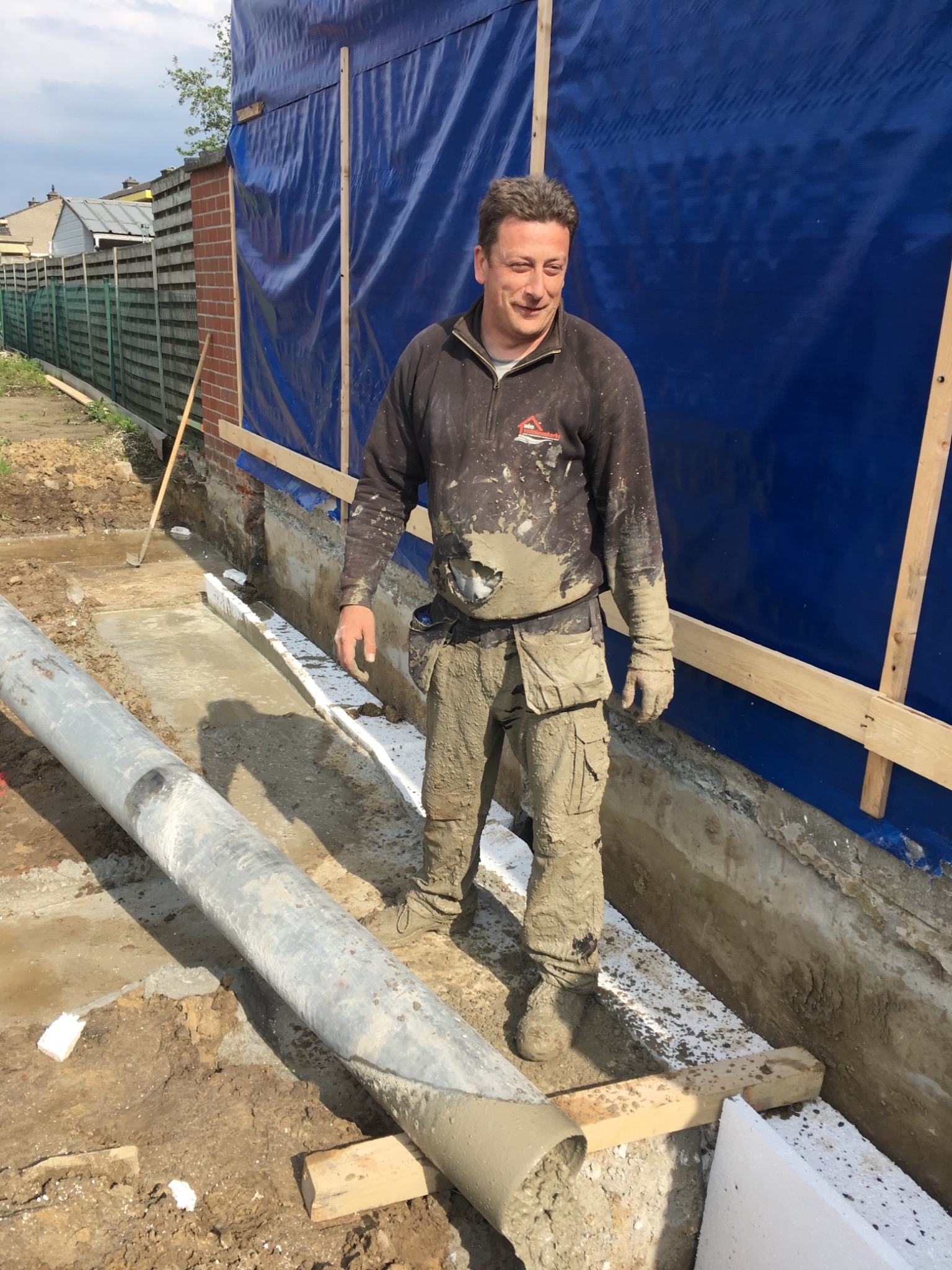 Uśmiechnięty pracownik budowlany, ubrudzony zaprawą, stoi w wykopie obok rury kanalizacyjnej i ocieplonych fundamentów budynku, zabezpieczonego niebieską folią.