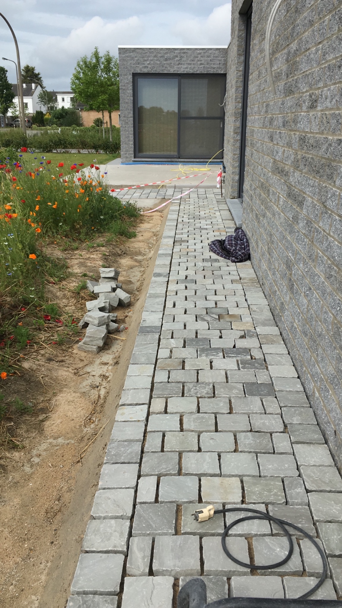 Układanie kostki brukowej z szarego kamienia przy ścianie budynku, widoczne narzędzia i materiały budowlane, kwiaty w tle.