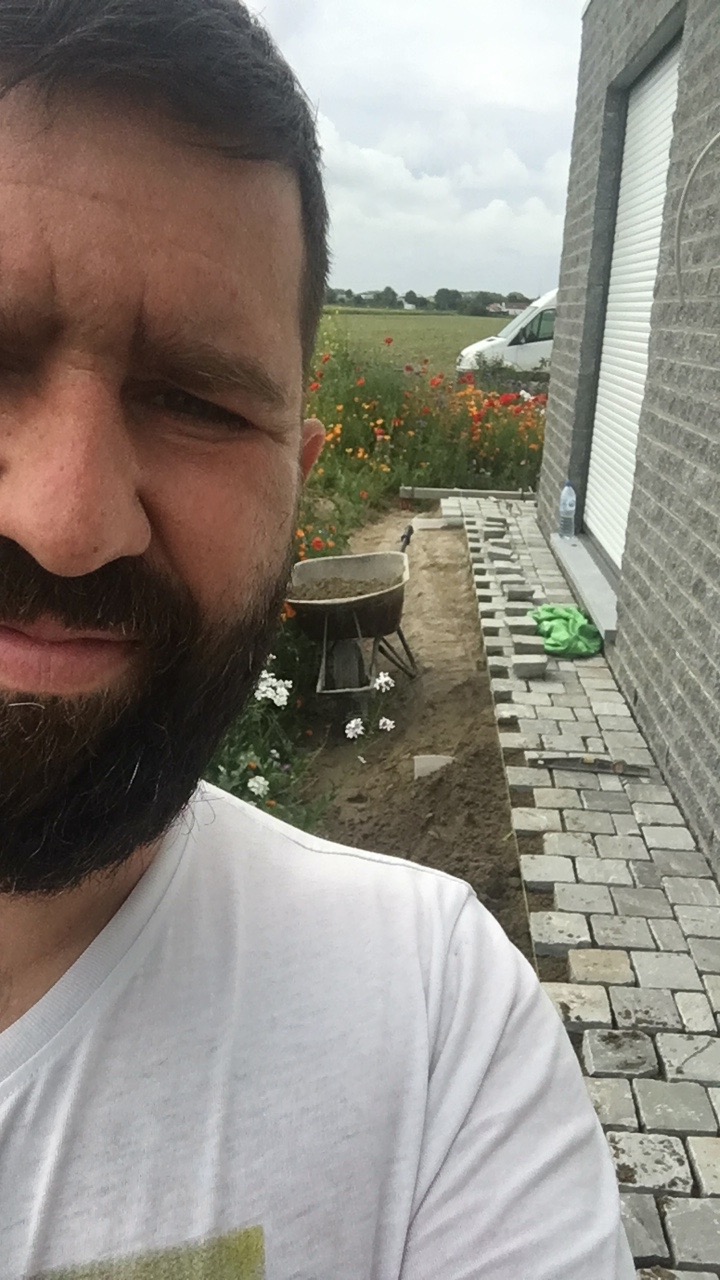 Mężczyzna robiący selfie na tle układanej ścieżki z kostki brukowej przy elewacji domu, widoczna taczka z ziemią, kwiaty i krajobraz polny w tle.