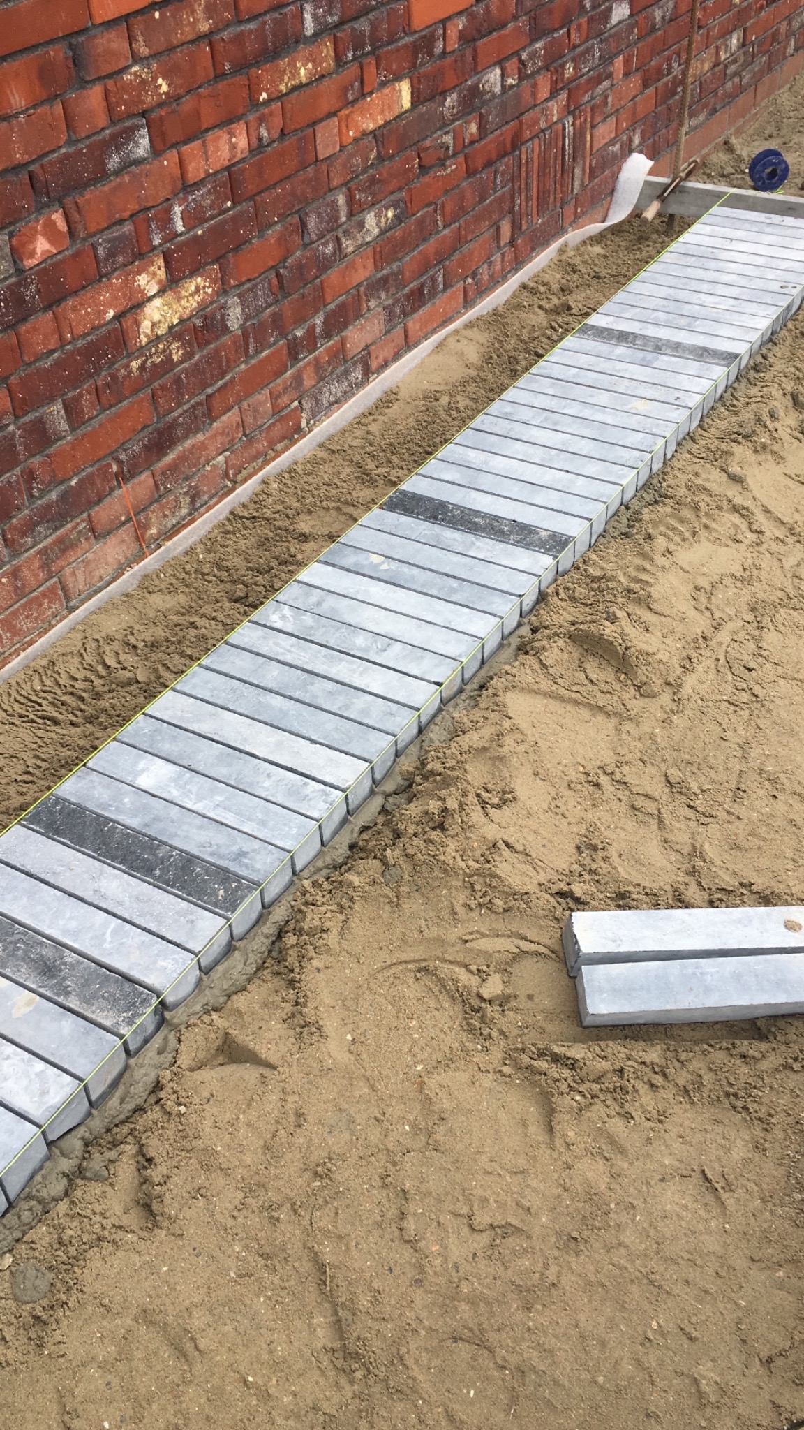 Układanie chodnika z szarych kostek brukowych wzdłuż ceglanej ściany, widoczne przygotowane podłoże z piasku i fragment linii wyznaczającej krawędź.