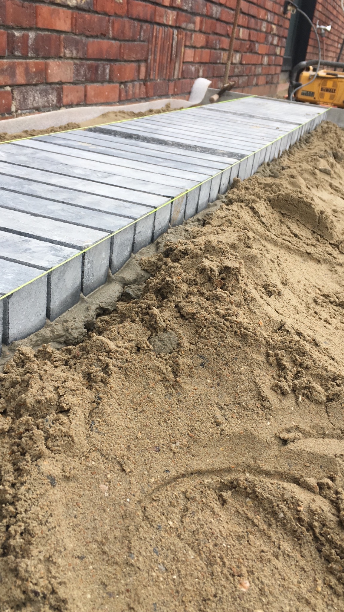 Układanie szarej kostki brukowej wzdłuż ceglanej ściany, widoczny piasek, zaprawa i żółta nić wyznaczająca linię.