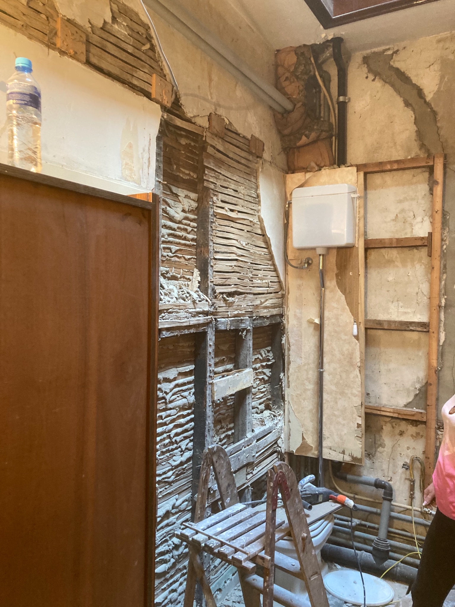Łazienka w trakcie demontażu starej zabudowy: odsłonięte drewniane elementy konstrukcyjne ściany, fragment instalacji wodnej i kanalizacyjnej, drabina malarska oraz butelka wody na półce.