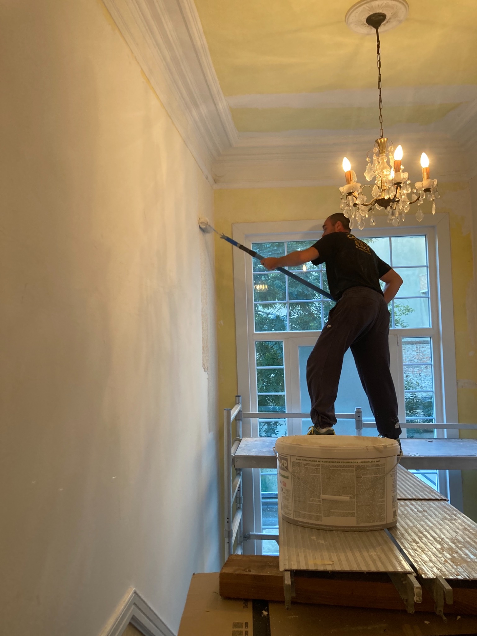 Mężczyzna maluje ścianę wałkiem na rusztowaniu w pokoju z kryształowym żyrandolem, widoczny kubeł z masą szpachlową.