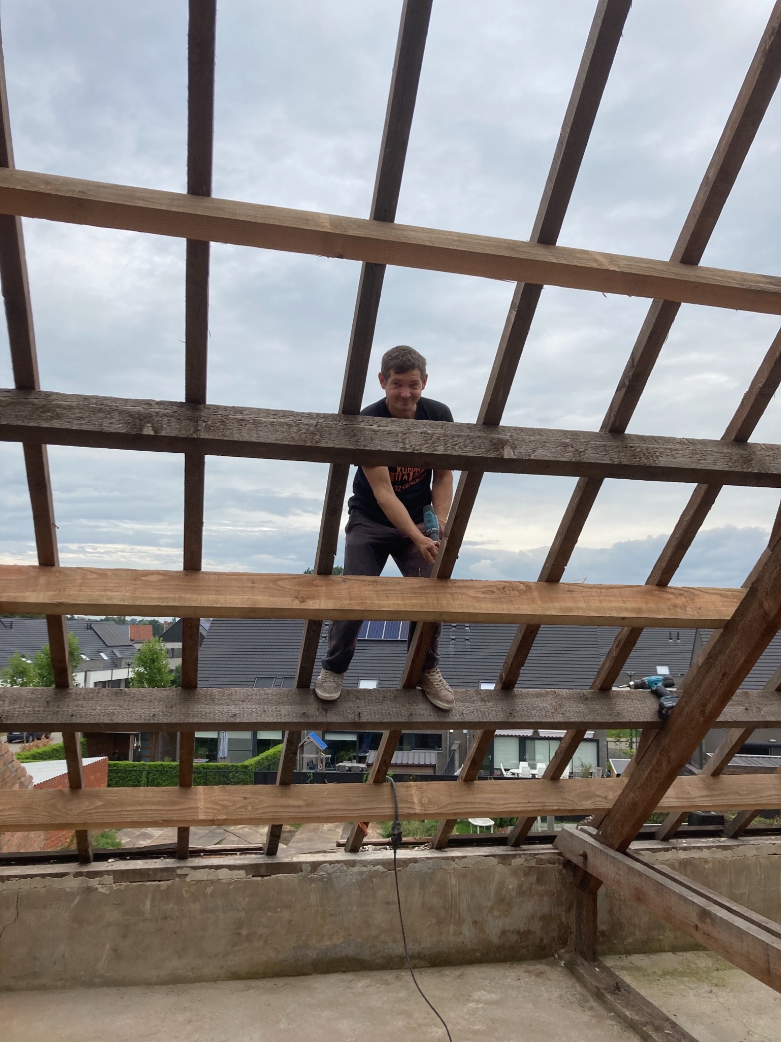 Pracownik z wiertarką na drewnianej konstrukcji dachu podczas prac na poddaszu, widok z dołu na tle pochmurnego nieba i zabudowań.