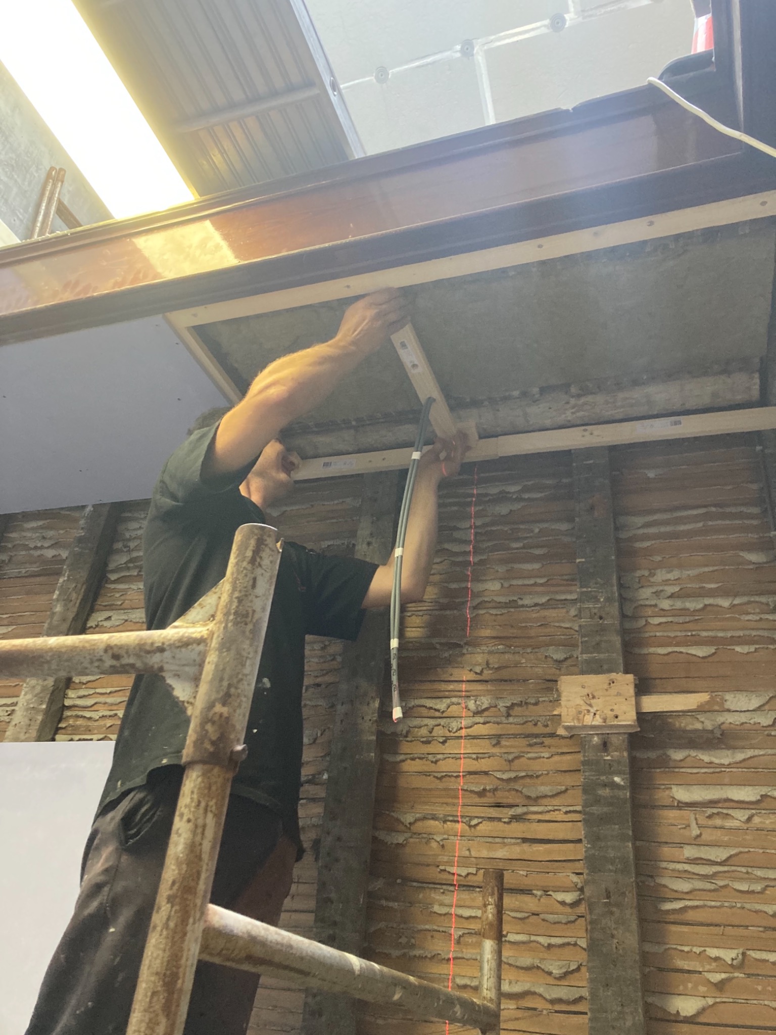 Pracownik na rusztowaniu montuje drewniane listwy do sufitu z widoczną instalacją elektryczną, ściany w stanie surowym z widocznymi warstwami starej farby i tynku.