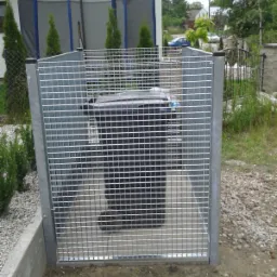 Metalowa kratownica zabezpieczająca pojemnik na śmieci, widoczna w ogrodzie przed domem.