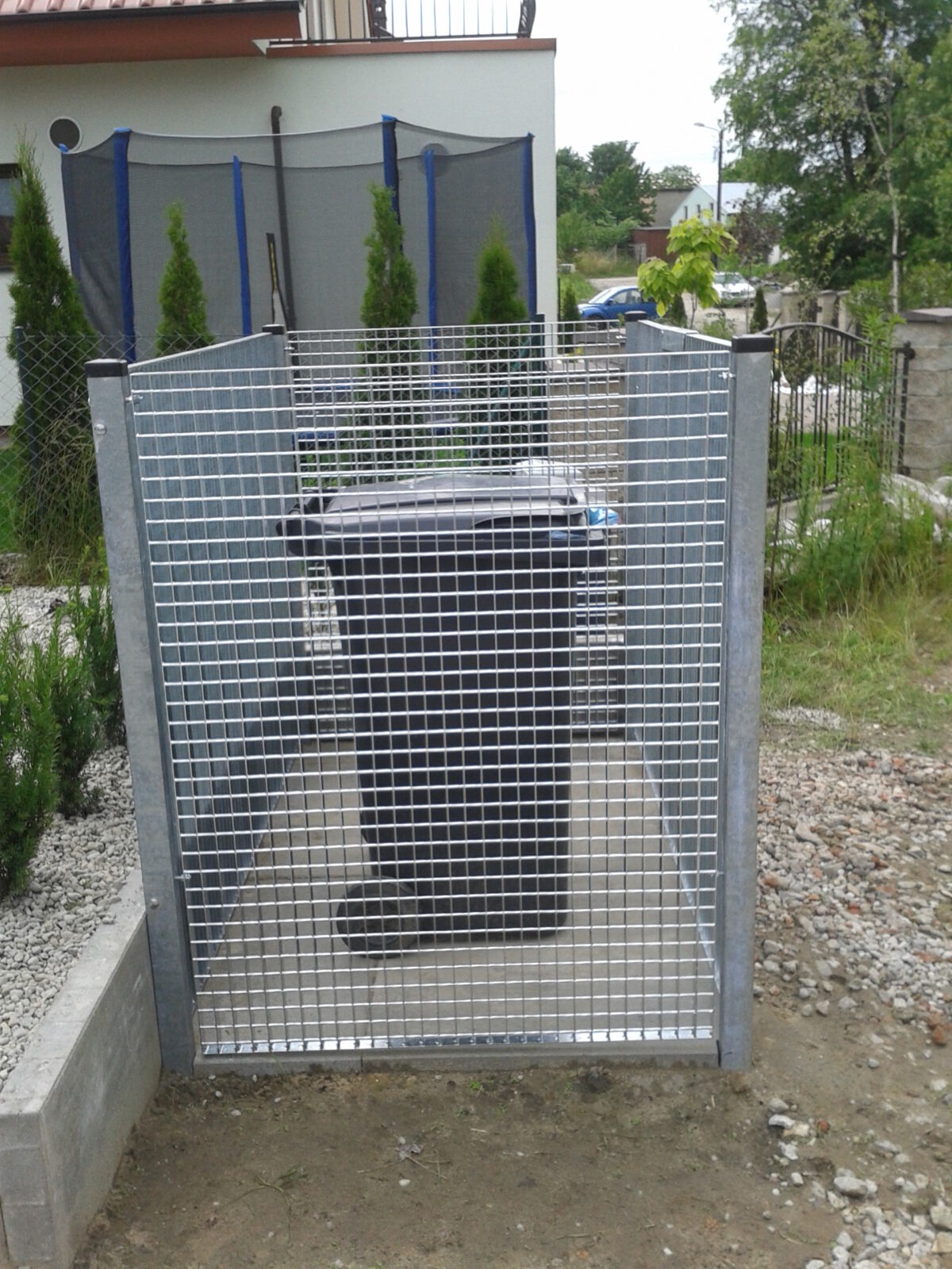 Metalowa kratownica zabezpieczająca pojemnik na śmieci, widoczna w ogrodzie przed domem.