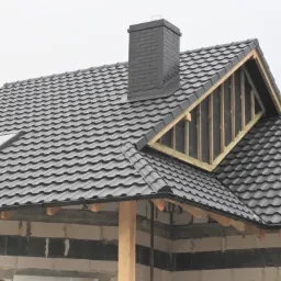 Nowy, ciemnoszary dach z dachówką ceramiczną i ceglanym kominem na surowym budynku, widoczne elementy konstrukcji drewnianej i okno dachowe.