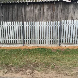 Nowoczesne ogrodzenie metalowe z szarymi słupkami i srebrnymi panelami pionowymi, osadzone na betonowej podmurówce, z brązową korą rozsypaną u podstawy, na tle starej drewnianej stodoły.