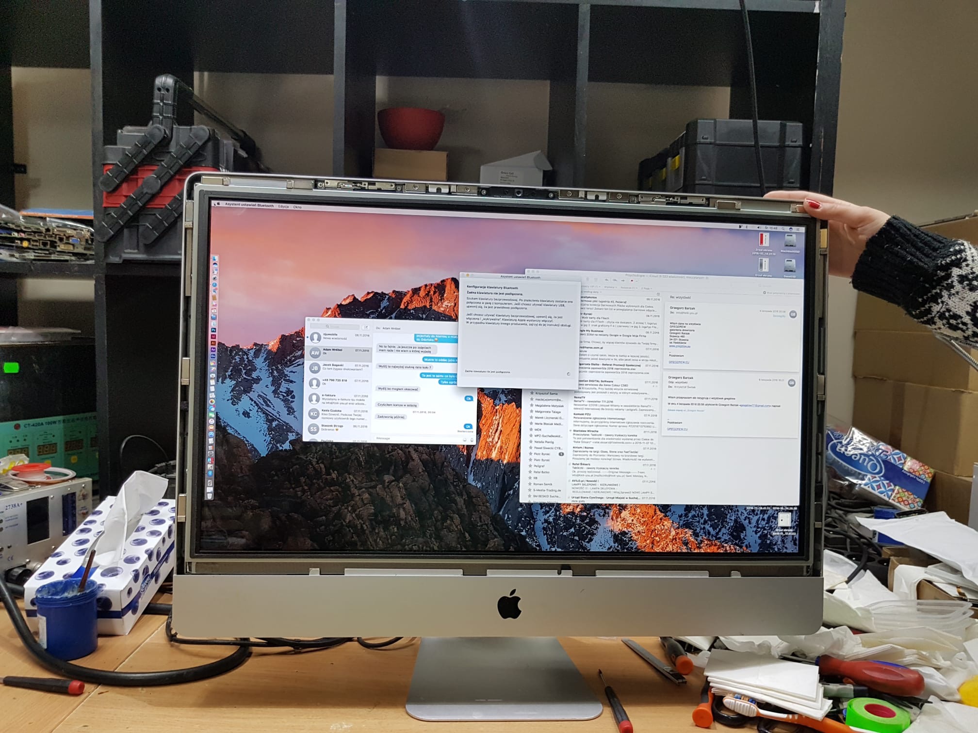 Otwarty iMac na biurku serwisowym z widocznymi narzędziami, otwartymi oknami poczty i ustawień Bluetooth, oraz dłonią kobiety trzymającą górną krawędź ekranu.