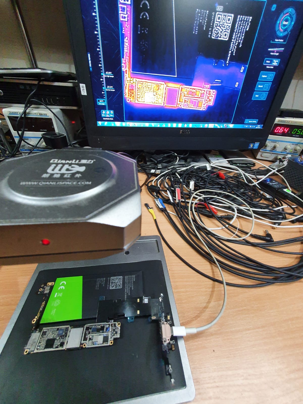 Płyta główna smartfona pod analizą termowizyjną, widoczna na ekranie monitora, otoczona narzędziami i przewodami na warsztacie.