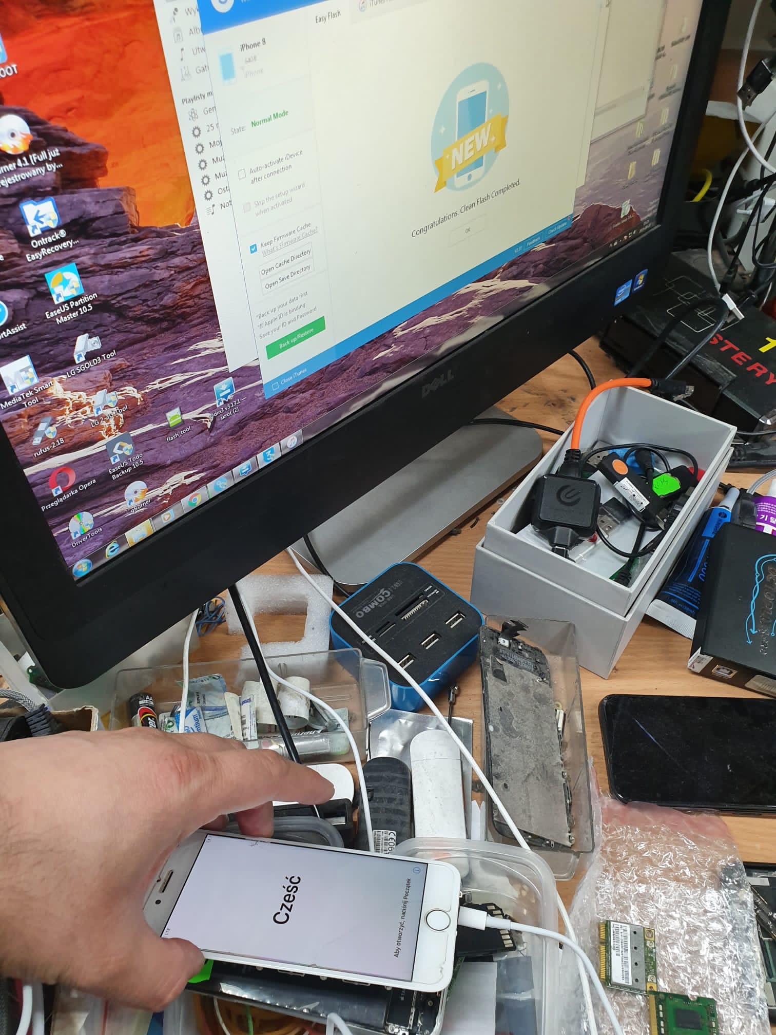 Technik podłącza iPhone'a z napisem 'Cześć' do komputera z uruchomionym oprogramowaniem do odzyskiwania danych; widoczne narzędzia i części zamienne na stole.