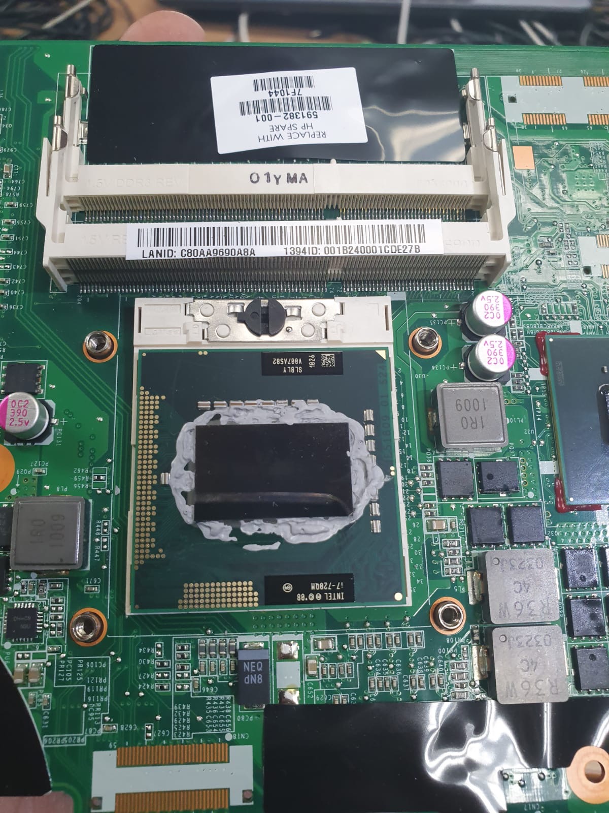 Płyta główna laptopa z zamontowanym procesorem Intel i7-720QM, pokrytym pastą termoprzewodzącą, widoczne gniazda pamięci RAM i liczne komponenty elektroniczne.