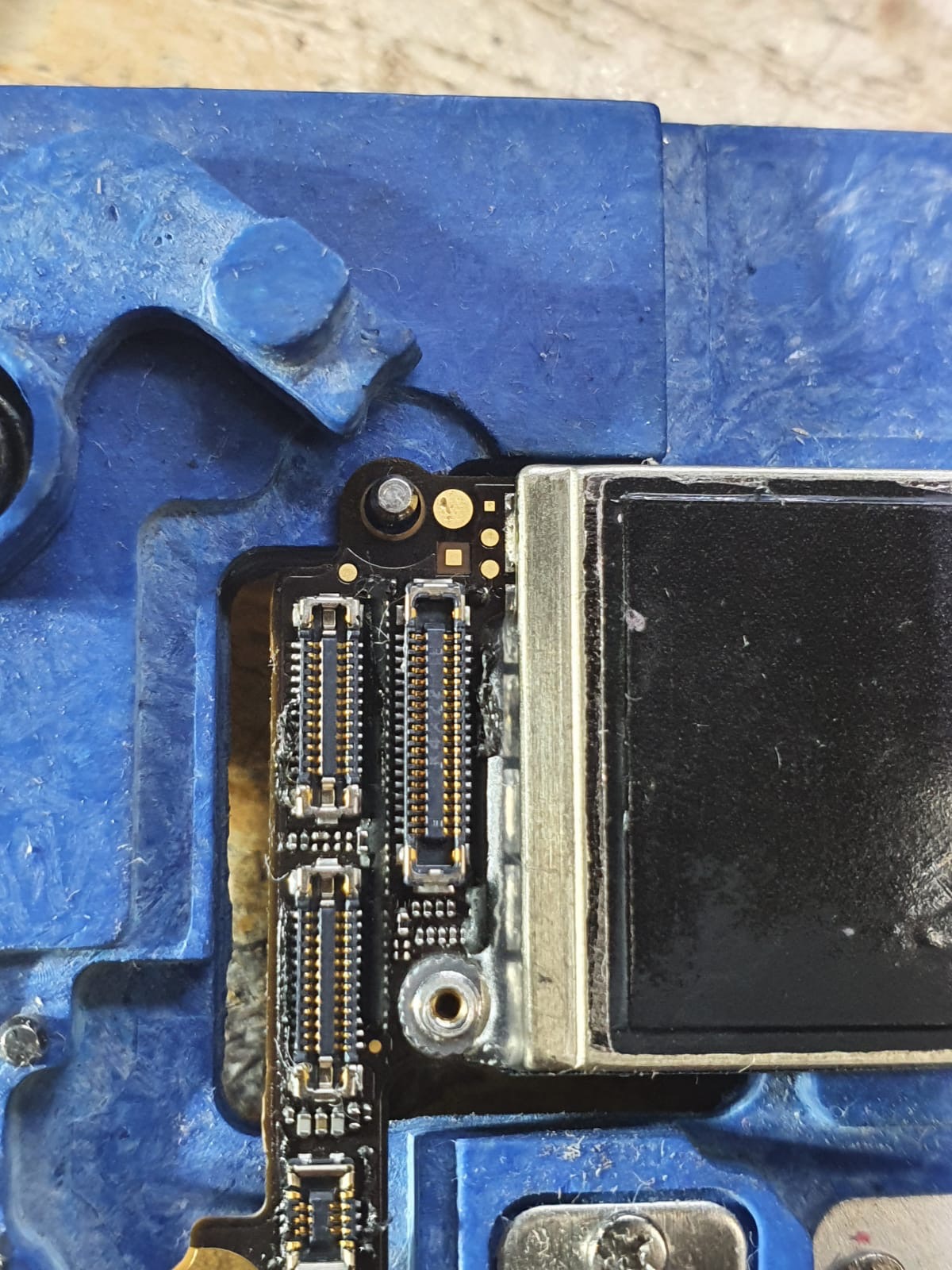 Fragment płyty głównej smartfona z widocznymi złączami, umieszczonej w niebieskim uchwycie montażowym.