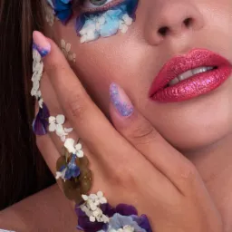 Portret kobiety z artystycznym makijażem z użyciem suszonych kwiatów na powiekach i dłoni, podkreślonymi brokatowymi cieniami i szminką.