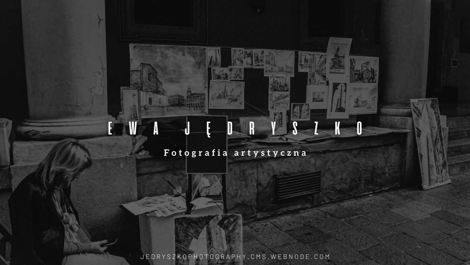 Czarno-białe zdjęcie przedstawia kobietę siedzącą na ławce, przeglądającą telefon. W tle kolumny, ściana z zawieszonymi szkicami architektonicznymi i obrazami. Na pierwszym planie widoczny stolik...