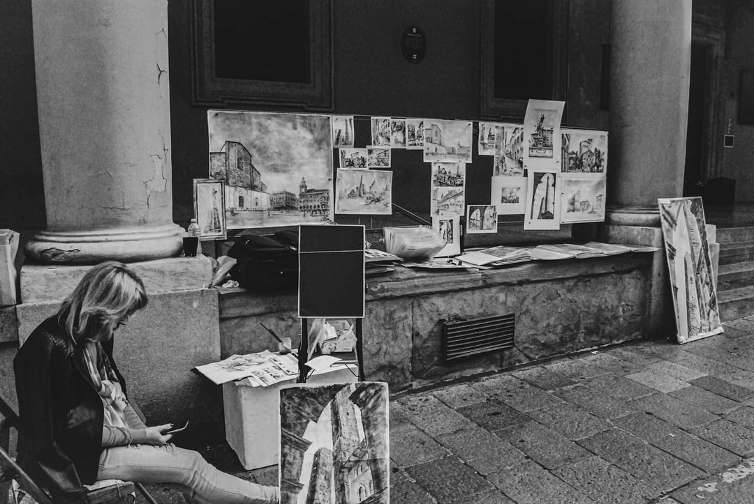 Czarno-białe zdjęcie przedstawia artystkę siedzącą na krześle i przeglądającą telefon, z wystawą jej szkiców architektonicznych i pejzaży miejskich umieszczonych na kamiennym murze za nią...