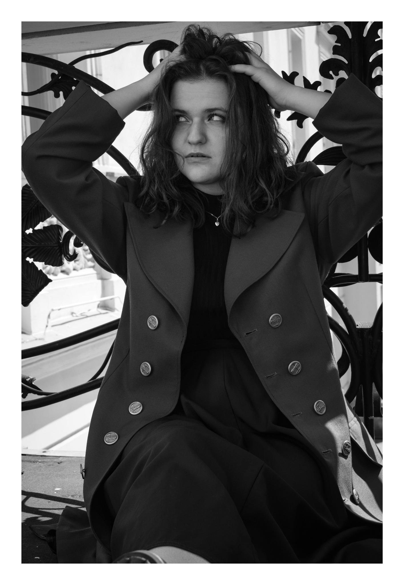 Czarno-białe zdjęcie portretowe kobiety w płaszczu, siedzącej na tle ozdobnej, żelaznej balustrady, z rękami uniesionymi do włosów i spoglądającej w bok.