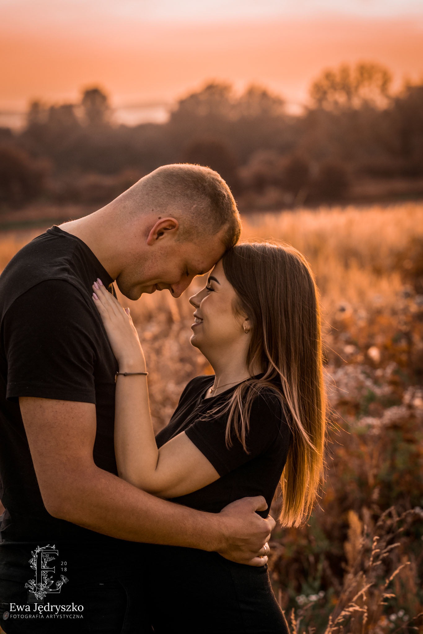 Zakochana para w objęciach na tle łąki o zachodzie słońca, ona uśmiecha się patrząc na niego.