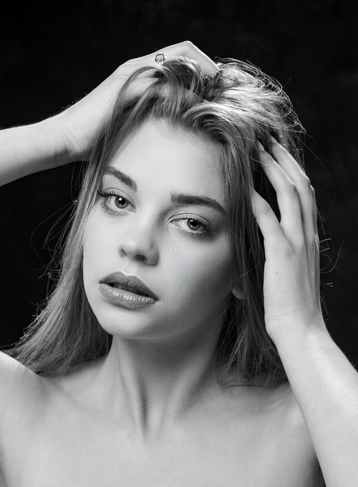 Portret kobiety w czerni i bieli, dłonie we włosach, pierścionek na palcu, intensywne spojrzenie w obiektyw.