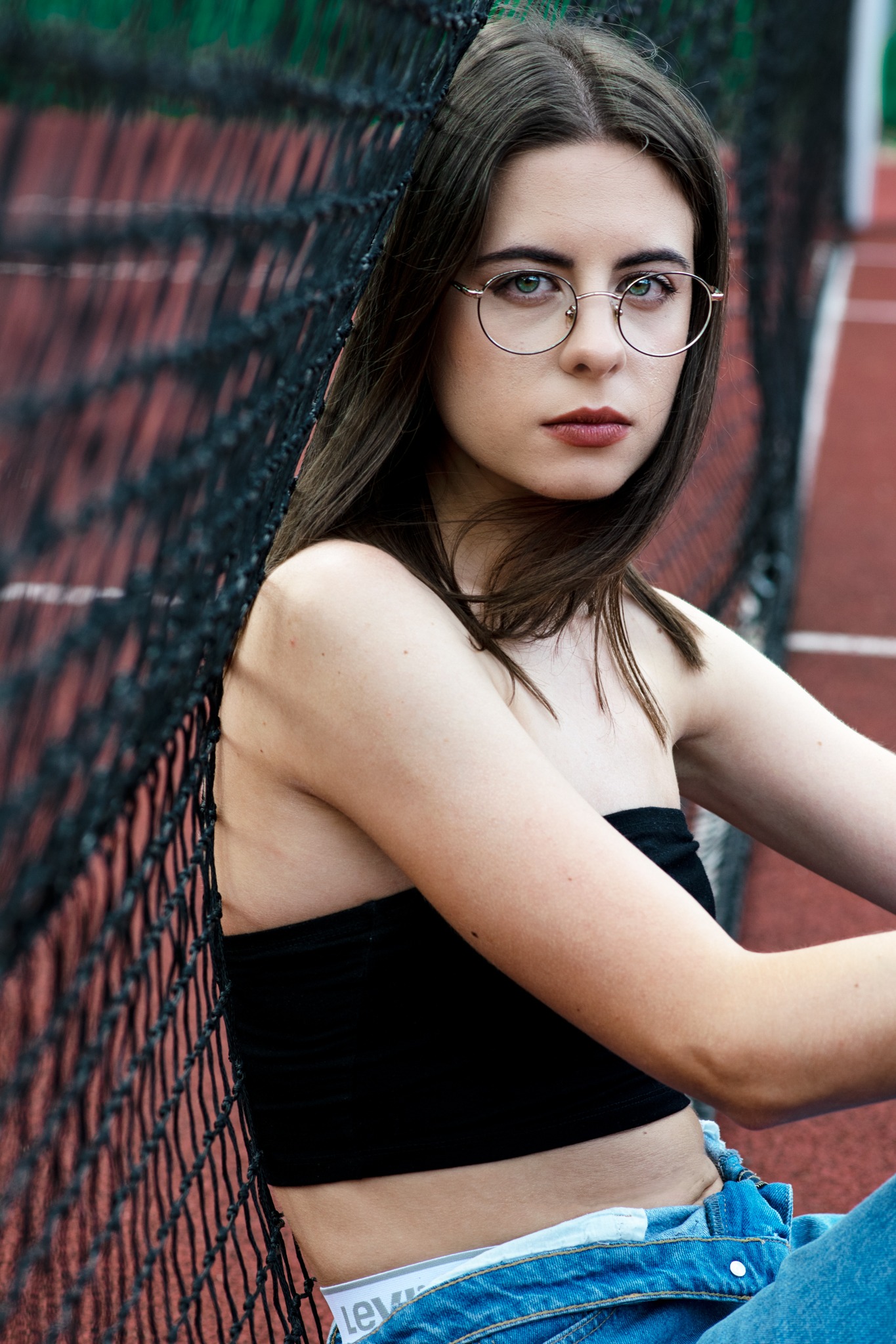 Portret młodej kobiety w okularach, opierającej się o siatkę na tle czerwonego kortu tenisowego.