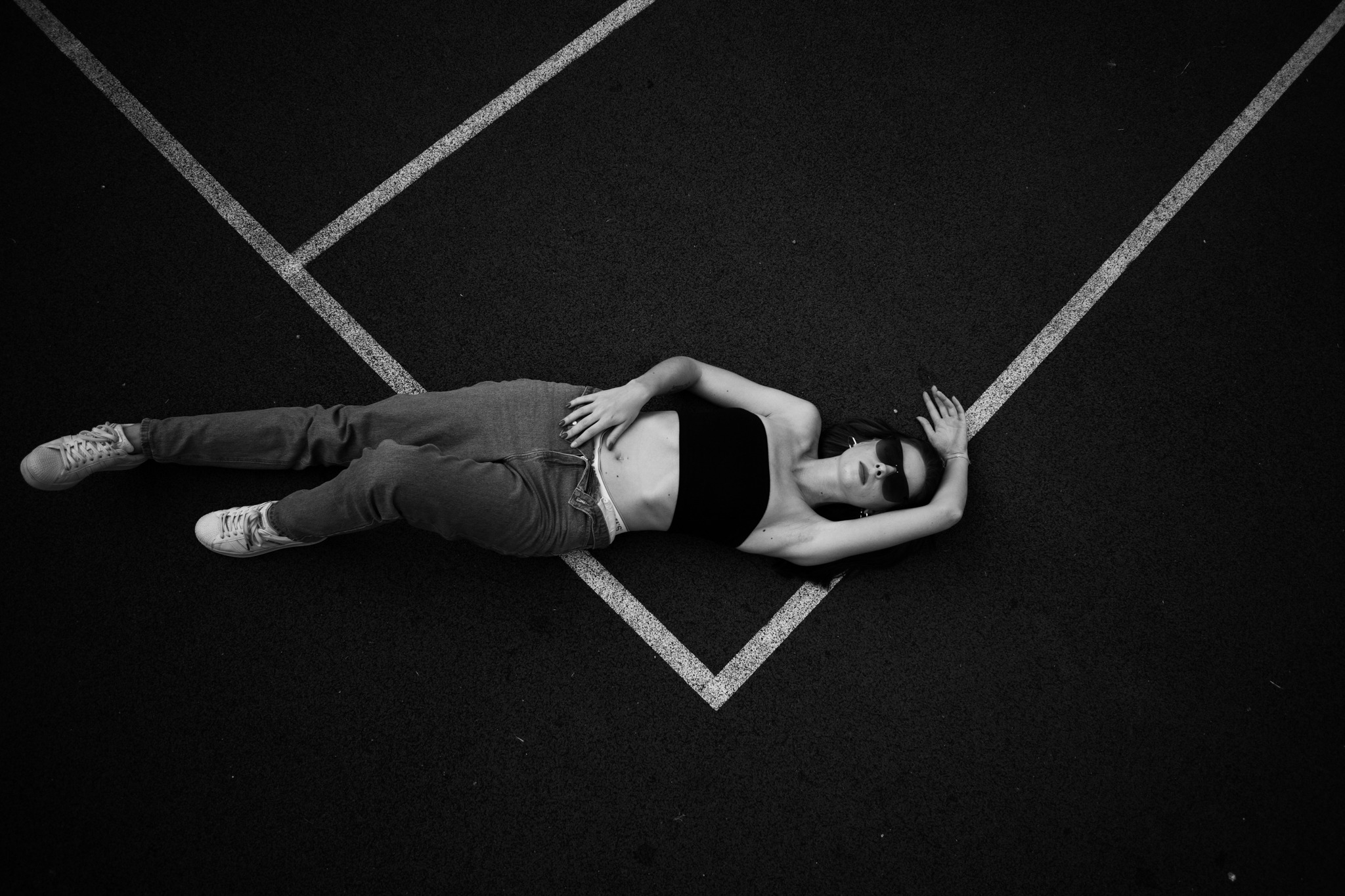 Czarno-białe zdjęcie z góry przedstawia kobietę w okularach przeciwsłonecznych, leżącą na czarnym podłożu z białymi liniami, ubrana w dżinsy i czarny top.