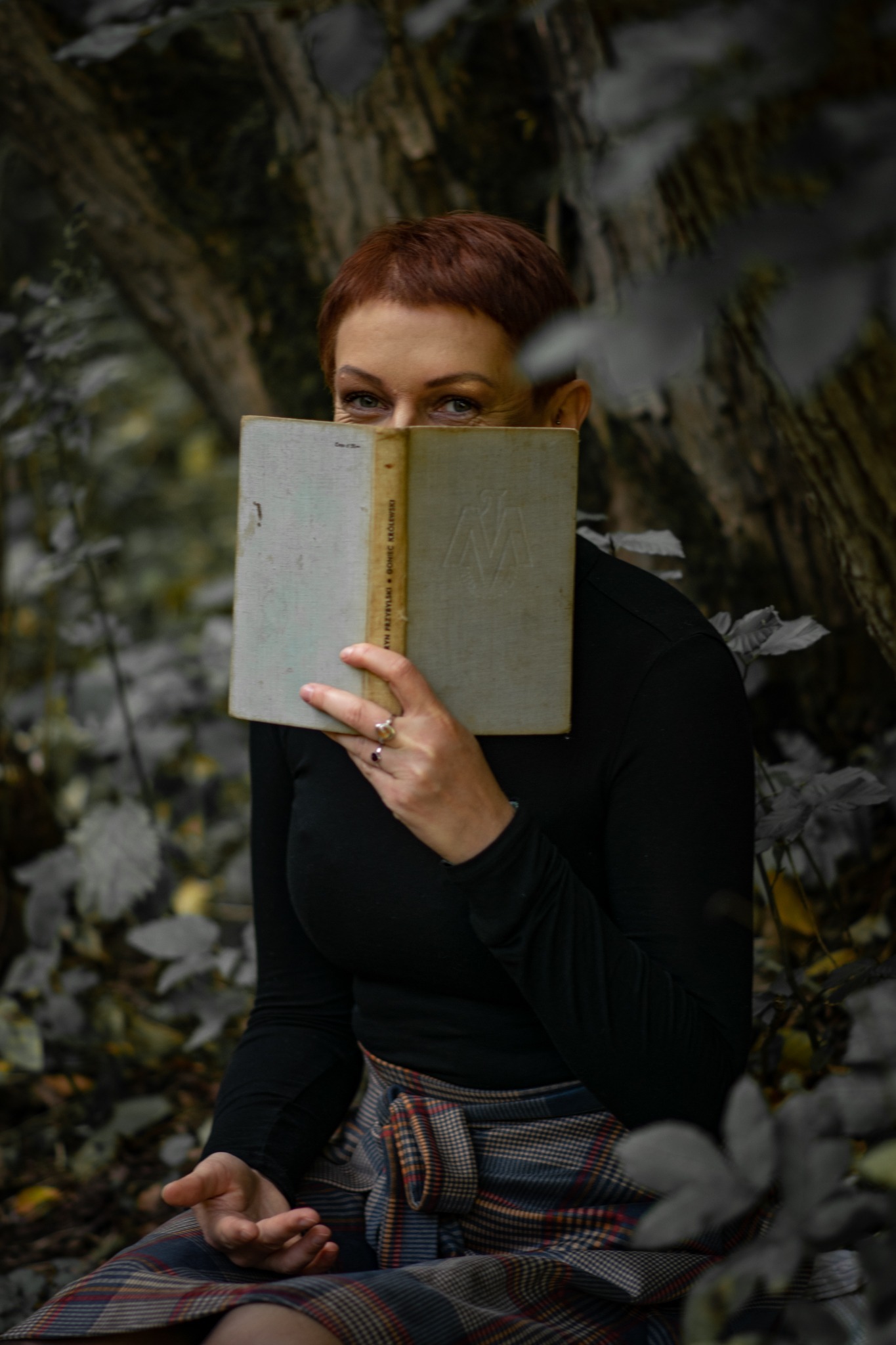 Kobieta z krótkimi rudymi włosami, siedząca na tle leśnym, zasłaniająca twarz starą książką z wytłoczonym monogramem na okładce, w czarnej bluzce i spódnicy w szkocką kratę.