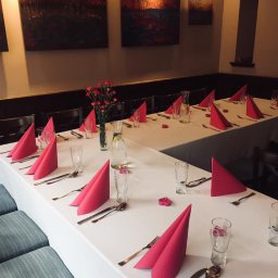 Restauracja U Artystów - Uroczysty stół z białym obrusem, nakryty dla gości z różowymi serwetkami, złotymi sztućcami i szklankami, ozdobiony wazonem z czerwonymi kwiatami, podświetlony ciepłym światłem, z obrazami...