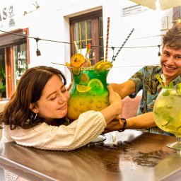 Restauracja U Artystów - Uśmiechnięta kobieta przytula ogromny, zielono-żółty koktajl z owocami, podczas gdy mężczyzna w kwiecistej koszuli trzyma drugi, mniejszy koktajl z limonkami i miętą, oboje siedzą przy drewnianym...