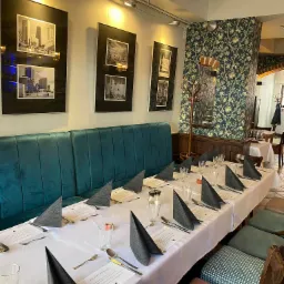 Długi stół nakryty białym obrusem z szarymi serwetkami ułożonymi w stożki, srebrnymi sztućcami i szklankami w restauracji z zielonymi tapicerowanymi krzesłami i kanapą oraz czarno-białymi zdjęciami...