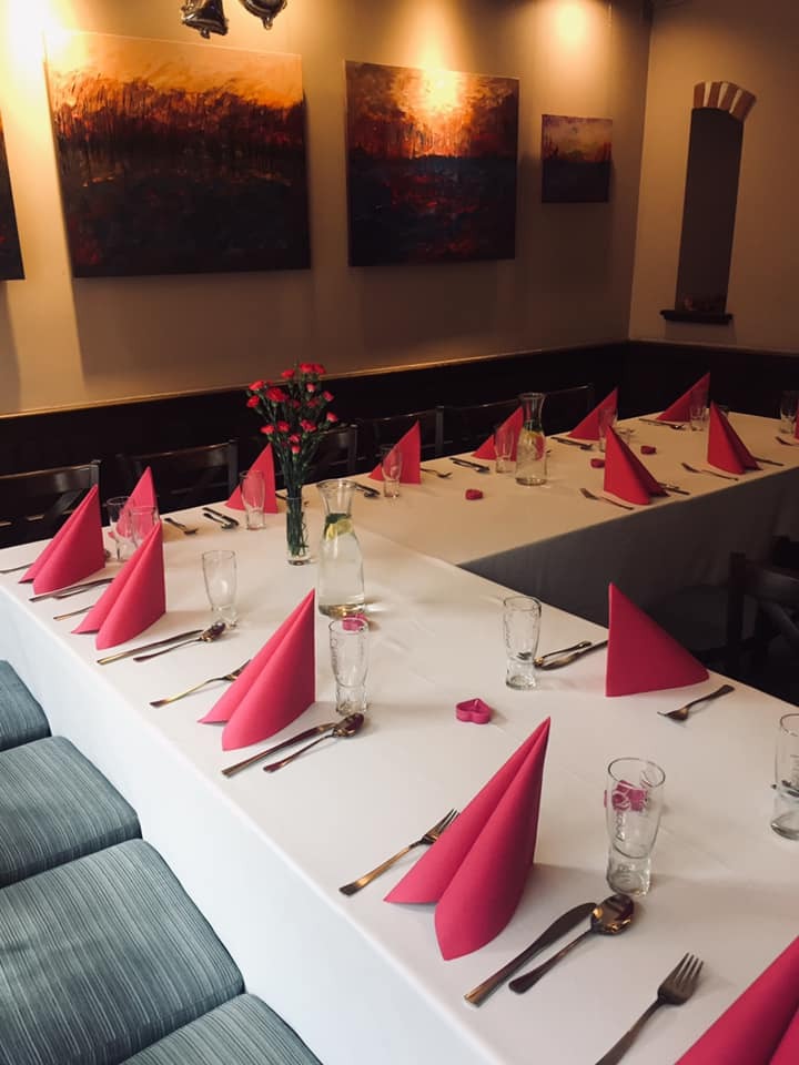 Uroczysty stół z białym obrusem, nakryty dla gości z różowymi serwetkami, złotymi sztućcami i szklankami, ozdobiony wazonem z czerwonymi kwiatami, podświetlony ciepłym światłem, z obrazami...