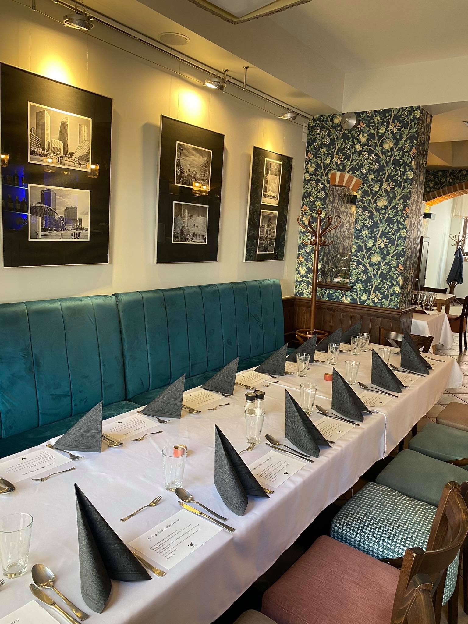 Długi stół nakryty białym obrusem z szarymi serwetkami ułożonymi w stożki, srebrnymi sztućcami i szklankami w restauracji z zielonymi tapicerowanymi krzesłami i kanapą oraz czarno-białymi zdjęciami...