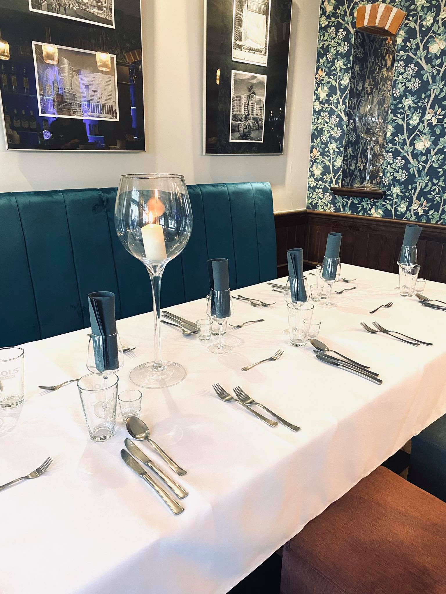Elegancko nakryty stół z białym obrusem, szkłem i sztućcami, z dekoracyjną świecą w dużym kielichu, ustawiony w restauracji z zieloną tapicerowaną ławą i wzorzystą tapetą.