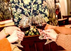 Dwie pary rąk wznoszą kieliszki z czerwonym winem w toaście, na tle wzorzystej tapety i ciepłego oświetlenia wnętrza.