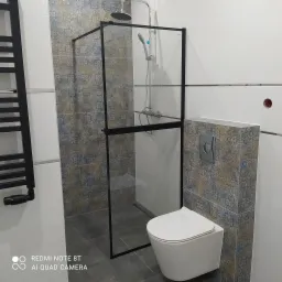 Nowoczesna łazienka z kabiną prysznicową typu walk-in z czarnymi profilami, wiszącą toaletą i grzejnikiem drabinkowym w kolorze czarnym, wykończona białymi płytkami oraz płytkami dekoracyjnymi...