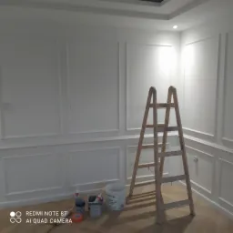 Białe ściany z dekoracyjnymi panelami, drewniana drabina, wiadro z farbą i akcesoria malarskie na podłodze z jasnego drewna, wnętrze w trakcie wykańczania.