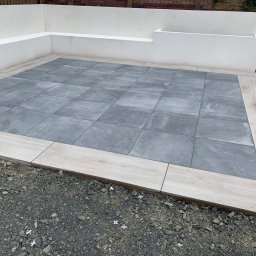 Dariusz Cholewa - Wykończony taras z szarych, kwadratowych płytek otoczonych jasnymi, drewnopodobnymi płytkami. Na obrzeżach tarasu widoczne drobne kamienie i krzyżyki dystansowe.