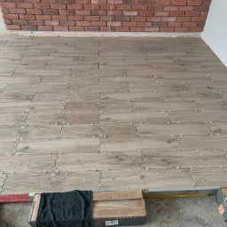 Ułożone płytki podłogowe imitujące drewno z krzyżykami dystansowymi, fragment ściany z cegły dekoracyjnej, poziomica i karton płytek na podłodze.