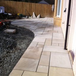 Dariusz Cholewa - Świeżo ułożony, beżowy chodnik z płyt kamiennych o nieregularnym kształcie, obok gruz i narzędzia budowlane w słoneczny dzień.