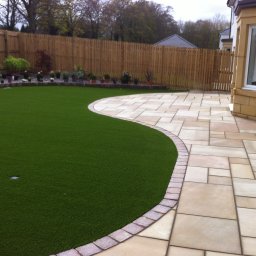 Dariusz Cholewa - Ułożona z nieregularnych, beżowych płyt ścieżka z kamienia graniczy z trawnikiem ze sztucznej trawy, oddzielonym obrzeżem z kostki; w tle drewniany płot.