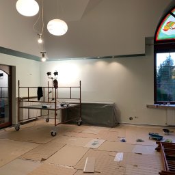 Dariusz Cholewa - Pokój w trakcie przygotowań do malowania: podłoga zabezpieczona kartonem, rusztowanie na kółkach, grzejnik osłonięty folią, widoczne narzędzia malarskie, okno z witrażem.