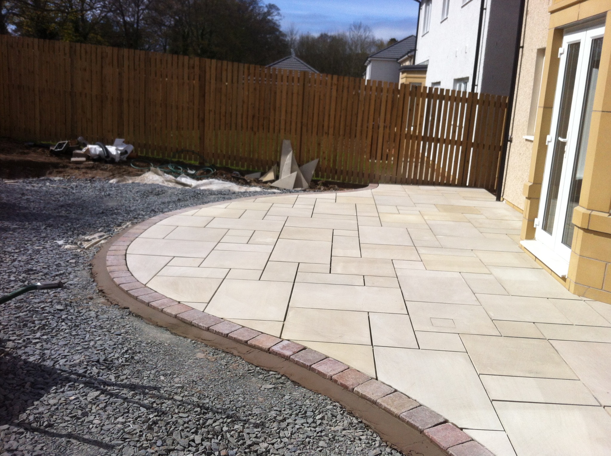 Ułożony z jasnych płyt taras z obrzeżem z czerwonej kostki, widok na drewniany płot i część domu z białymi drzwiami.