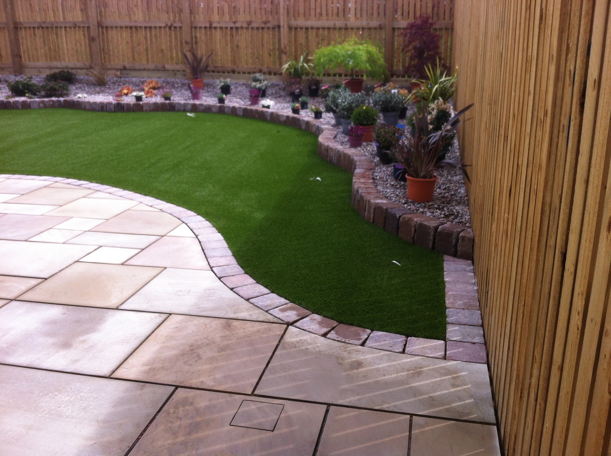 Ułożona z jasnych płyt ścieżka na zewnątrz domu, przylegająca do sztucznej trawy i rabaty z roślinami w doniczkach, oddzielonej obrzeżem z kamienia, z drewnianym płotem w tle.