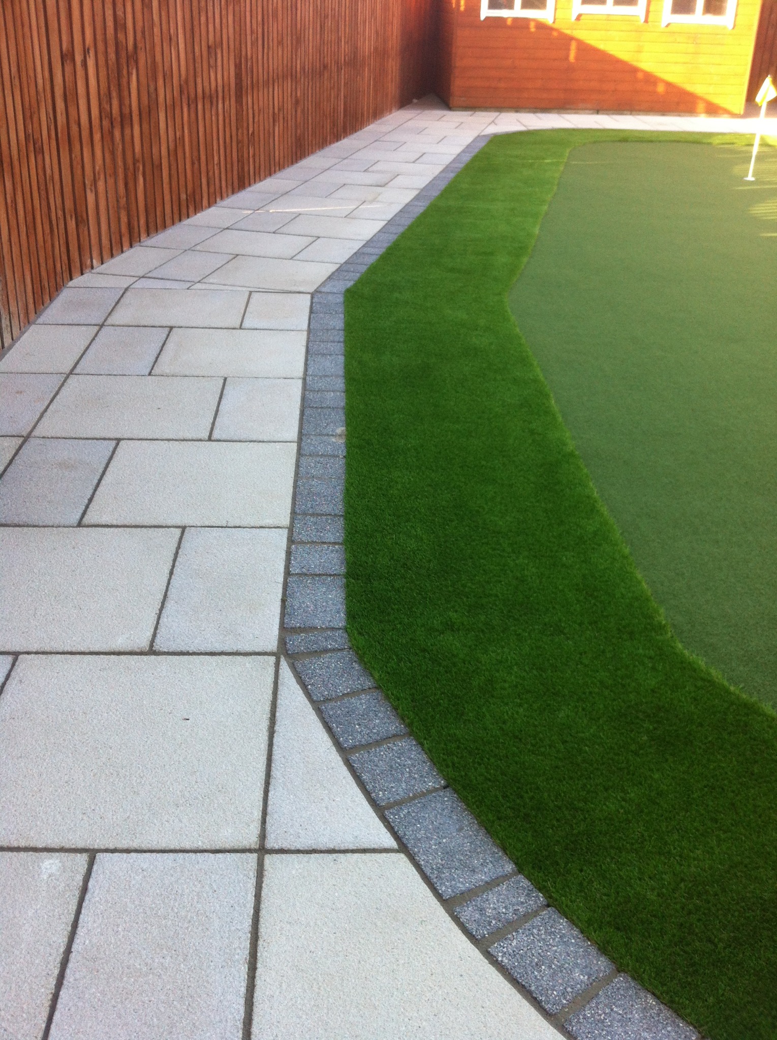 Ułożona z jasnych płyt ścieżka obok drewnianego ogrodzenia i sztucznej trawy, z widocznym fragmentem budynku w tle.