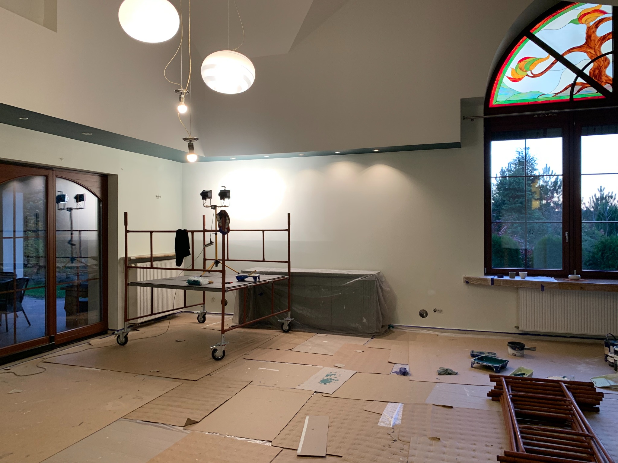 Pokój w trakcie przygotowań do malowania: podłoga zabezpieczona kartonem, rusztowanie na kółkach, grzejnik osłonięty folią, widoczne narzędzia malarskie, okno z witrażem.