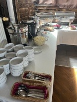 Stół cateringowy z białym obrusem, na którym znajdują się białe miseczki, koszyczki ze srebrnymi łyżkami, pojemniki do podgrzewania potraw oraz inne naczynia i jedzenie. Tło stanowią drewniane panele.