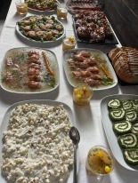 Stół z różnorodnymi potrawami: sałatka jarzynowa z łyżką, galaretki z krewetkami, roladki szpinakowe, tatar, przystawki w słoiczkach. Biały obrus, ciemne tło.