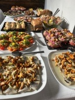 Stół z różnorodnymi potrawami: śledzie z kurkami i cebulą, sałatki warzywne, przystawki z pomidorami i ziołami, pieczywo. Ułożone na białych talerzach i czarnych deskach, tworzą bogaty bufet.